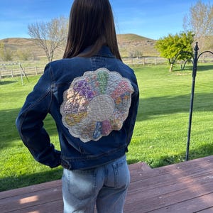 May include: A dark blue denim jacket with a colorful, circular quilt patch on the back. The patch features a variety of pastel colors and a cream-colored center. The jacket is paired with light blue jeans.