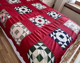 May include: A handmade quilt with a deep red background and geometric patchwork squares. The squares feature various patterns in white, blue, green, and black. The quilt is displayed on a white metal bed frame, showcasing a vintage aesthetic.