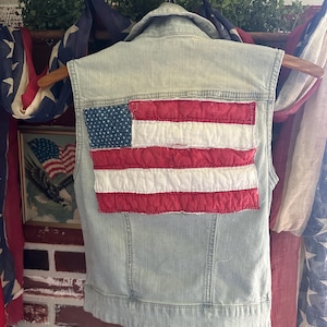 May include: Light blue denim vest with an American flag patch on the back. The flag features red and white stripes and a blue field with white stars. The vest is hanging on a wooden hanger.