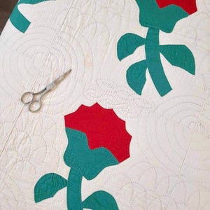 May include: A white quilt with a repeating pattern of red and green flowers. The quilt is hand-stitched and has a vintage look.