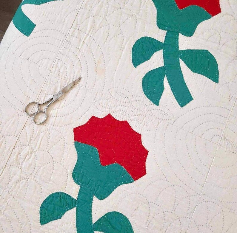 May include: A white quilt with a repeating pattern of red and green flowers. The quilt is hand-stitched and has a vintage look.