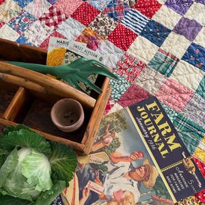May include: A wooden garden caddy containing a green hand rake, a small terracotta pot, and two seed packets for Marigold and Sweet Alyssum. Beside it lies a January 1936 Farm Journal magazine and a head of green cabbage on a patchwork quilt.