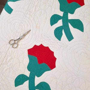 May include: A white quilt with a red and green floral pattern. The quilt is quilted with a circular pattern. A pair of silver scissors is lying on the quilt.