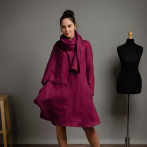 May include: A deep magenta long-sleeved linen dress with a matching fringed scarf. The dress features a loose, flared silhouette that falls above the knee, long sleeves, and a simple round neckline. The fabric has a visible, textured weave.