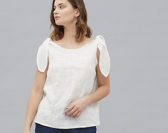 Puede incluir: Una blusa de lino blanca con tirantes de lazo y cuello redondo. La modelo lleva pantalones cortos de lino azul marino y sandalias de tacón negras.