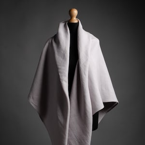 Puede incluir: Un chal de lino gris claro drapeado sobre un maniquí. El chal está hecho de una tela suave y natural y tiene un diseño simple y elegante.