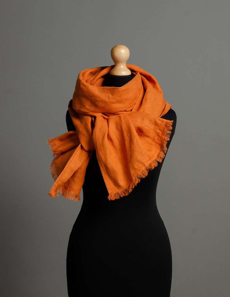 May include: A burnt orange linen scarf with frayed edges, draped around a black mannequin.