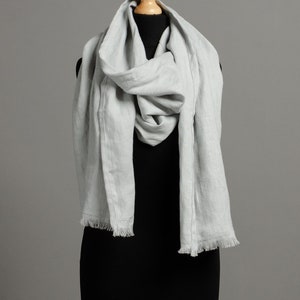 May include: A light gray linen scarf with a fringed edge. The scarf is draped around a black mannequin.