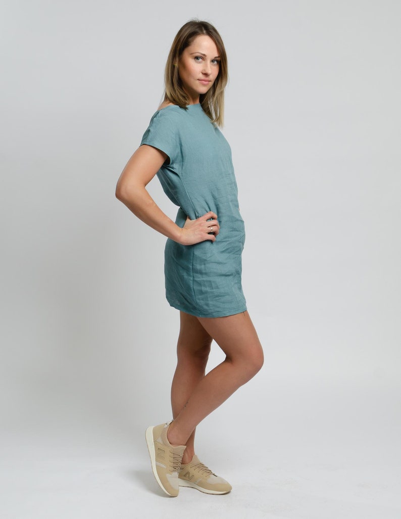 Op de afbeelding: Een vrouw in een turquoise linnen jurk en beige sneakers. Ze staat met haar hand op haar heup en kijkt naar de camera.