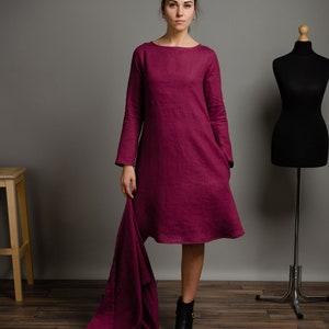 May include: A knee-length magenta dress with long sleeves, a round neckline, and a loose, A-line silhouette. The garment features a textured fabric finish and includes a matching magenta fabric piece draped from the side, with LinenStitchStore &copy; All Rights Reserved text visible at the bottom right.