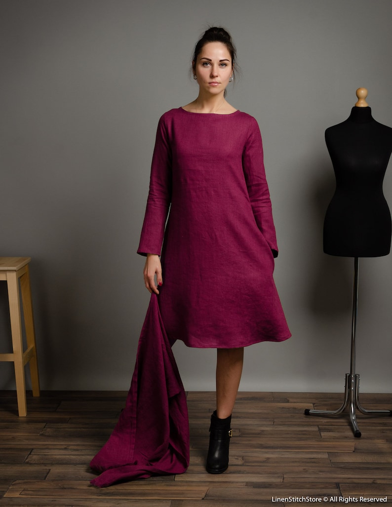 May include: A knee-length magenta dress with long sleeves, a round neckline, and a loose, A-line silhouette. The garment features a textured fabric finish and includes a matching magenta fabric piece draped from the side, with LinenStitchStore &copy; All Rights Reserved text visible at the bottom right.