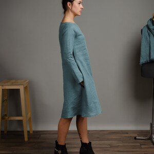 May include: A teal linen dress with long sleeves and a loose fit. The dress is shown on a model wearing black ankle boots.