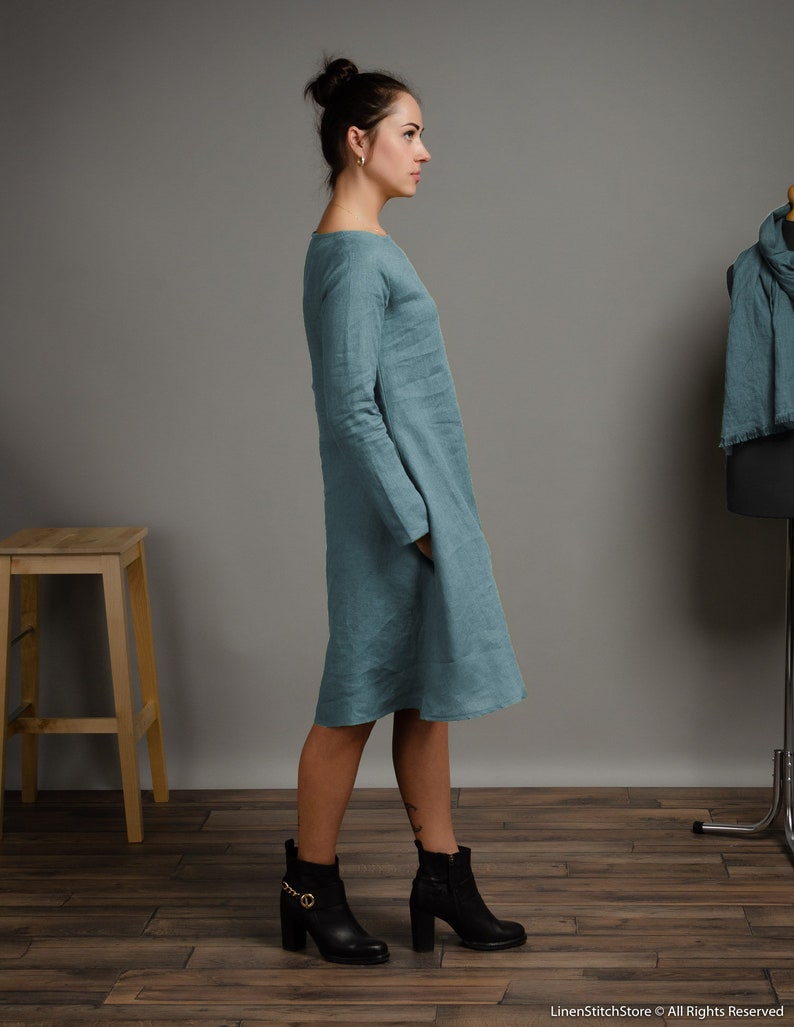 May include: A teal linen dress with long sleeves and a loose fit. The dress is shown on a model wearing black ankle boots.