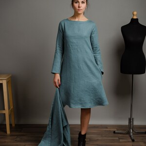 May include: A teal linen dress with long sleeves and a loose fit. The dress has a scoop neckline and a slightly flared skirt. The model is wearing black ankle boots.