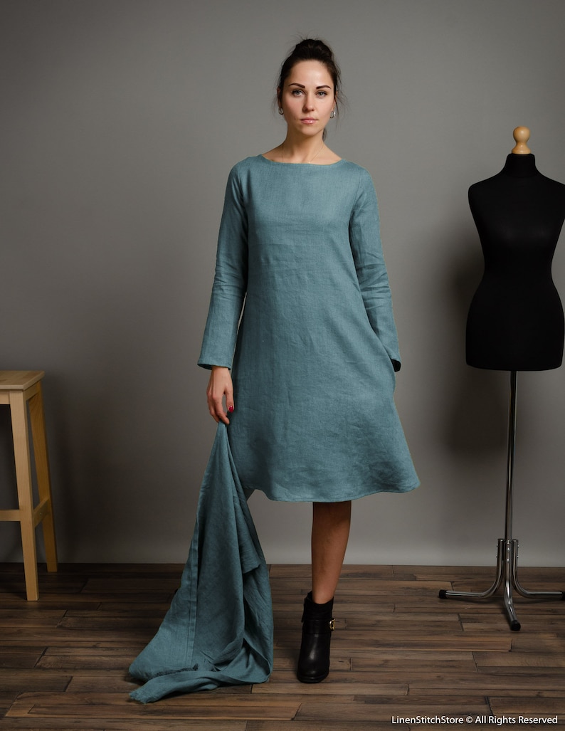 May include: A teal linen dress with long sleeves and a loose fit. The dress has a scoop neckline and a slightly flared skirt. The model is wearing black ankle boots.