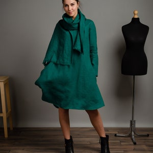 May include: A woman wearing a green linen dress with a matching green linen scarf. She is wearing black ankle boots and standing on a wooden floor.
