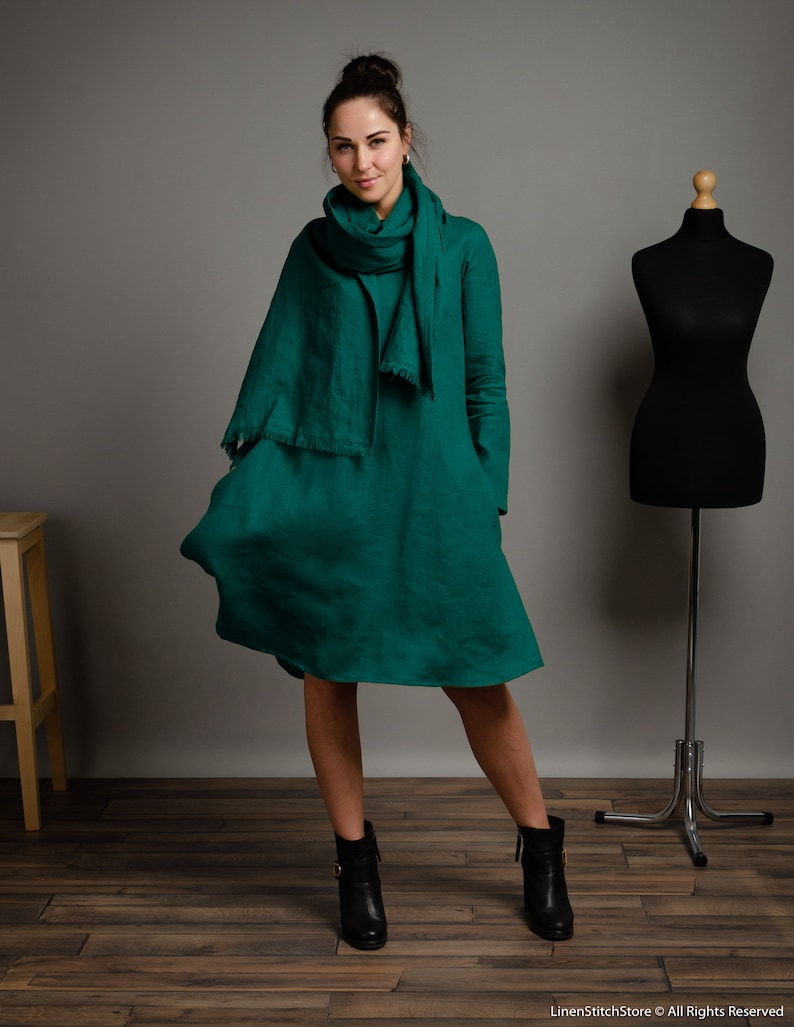 May include: A woman wearing a green linen dress with a matching green linen scarf. She is wearing black ankle boots and standing on a wooden floor.