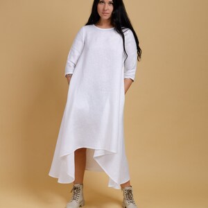 May include: A white linen dress with a loose fit and a high-low hemline. The dress has a round neckline and three-quarter sleeves. The model is wearing the dress with beige ankle boots.