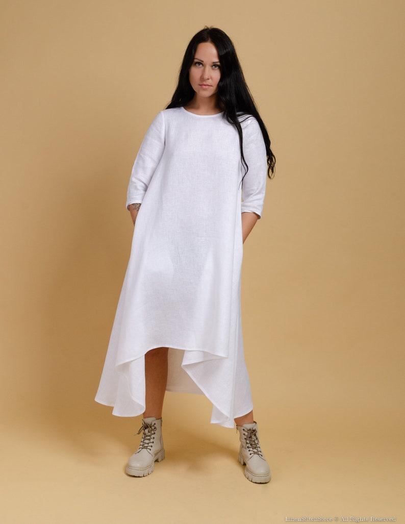 May include: A white linen dress with a loose fit and a high-low hemline. The dress has a round neckline and three-quarter sleeves. The model is wearing the dress with beige ankle boots.
