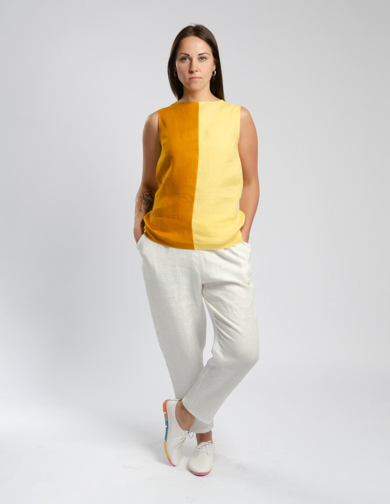 May include: A woman is wearing a sleeveless top with a yellow and orange color block design. She is wearing white pants and white shoes with a rainbow stripe on the sole.