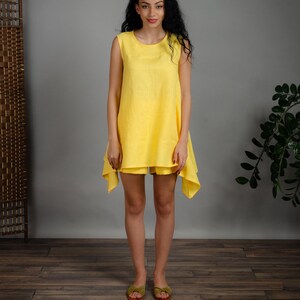 May include: A sleeveless yellow linen dress with a loose fit and a slightly asymmetrical hemline. The dress is worn by a model with long dark hair and is standing on a wooden floor.