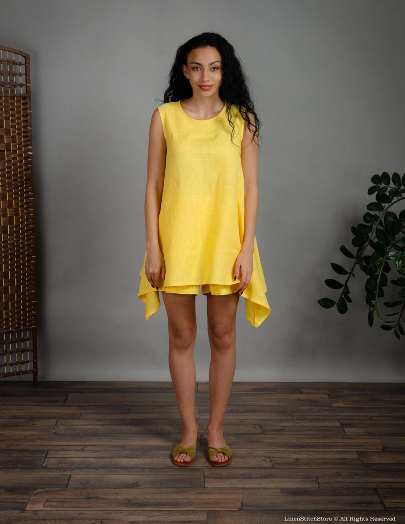 May include: A sleeveless yellow linen dress with a loose fit and a slightly asymmetrical hemline. The dress is worn by a model with long dark hair and is standing on a wooden floor.