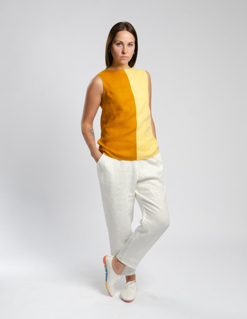 May include: A woman wearing a sleeveless top with a yellow and orange color block design. The top has a scoop neck and a relaxed fit. She is wearing white linen pants and white sneakers with a rainbow stripe.