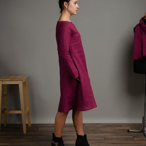 May include: A magenta knee-length dress with long sleeves and a round neckline. The garment features a straight silhouette with side pockets and a visible textured fabric finish. The dress is shown from a side profile view, highlighting the cut.