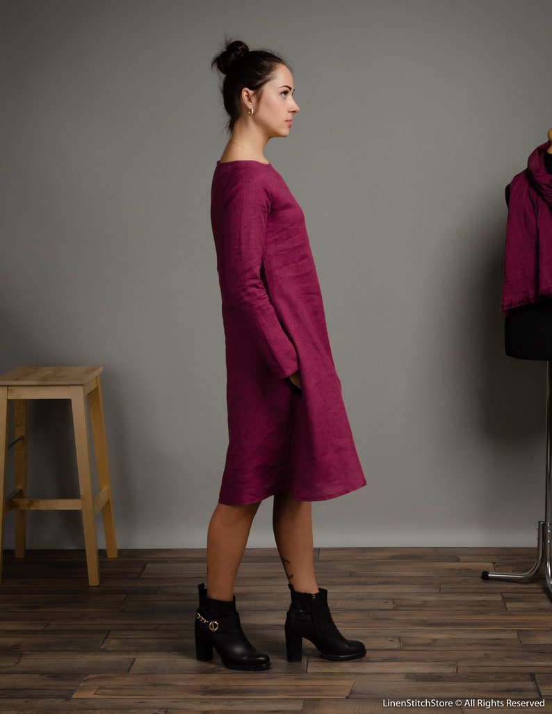 May include: A magenta knee-length dress with long sleeves and a round neckline. The garment features a straight silhouette with side pockets and a visible textured fabric finish. The dress is shown from a side profile view, highlighting the cut.