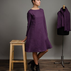 May include: A woman wearing a long-sleeved, purple linen dress with a boat neck. She is standing on a wooden floor with a wooden stool and a metal stand with a purple scarf draped over it.