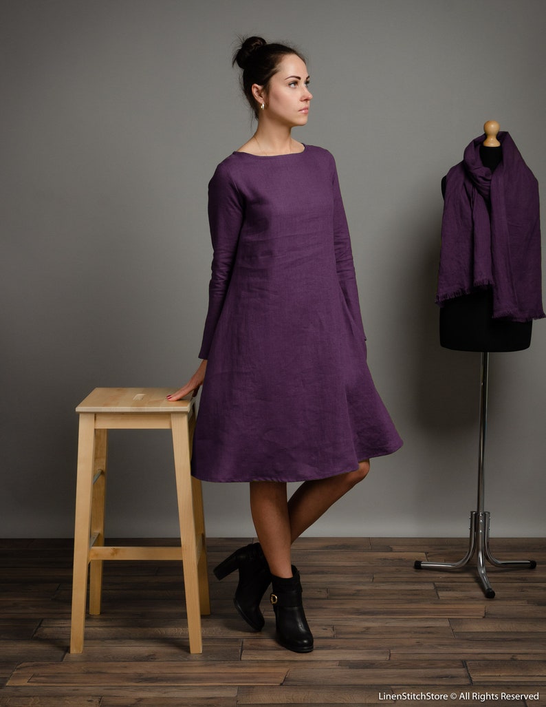 May include: A woman wearing a long-sleeved, purple linen dress with a boat neck. She is standing on a wooden floor with a wooden stool and a metal stand with a purple scarf draped over it.