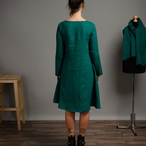 May include: A long-sleeved, emerald green linen dress with a scoop neck and a flowing skirt. The dress is shown on a model with tattoos on her ankles.