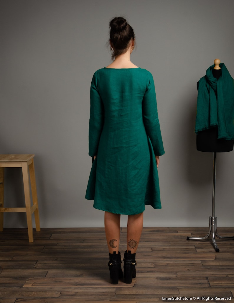 May include: A long-sleeved, emerald green linen dress with a scoop neck and a flowing skirt. The dress is shown on a model with tattoos on her ankles.