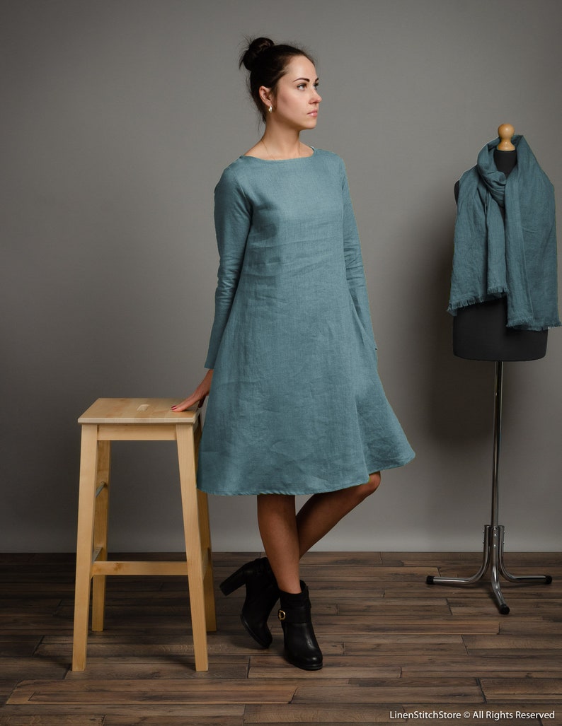 May include: A teal linen dress with a boat neck and 3/4 sleeves. The dress is loose fitting and falls to the knees. The model is wearing black ankle boots.