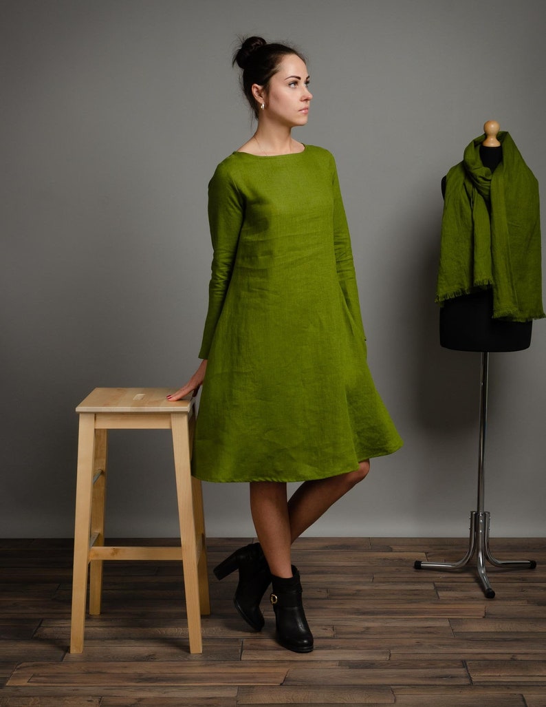 May include: A woman wearing a green linen dress with long sleeves and a loose fit. She is standing on a wooden floor and leaning against a wooden stool. She is wearing black ankle boots.