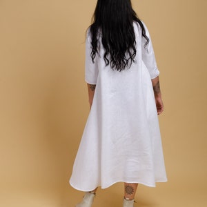 May include: A white linen dress with a long, flowing skirt and a simple, collared neckline. The dress is worn by a person with long, black hair.