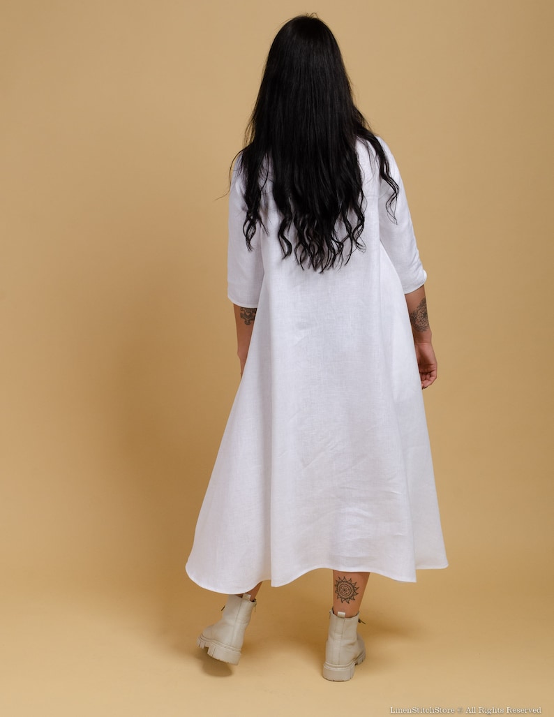 May include: A white linen dress with a long, flowing skirt and a simple, collared neckline. The dress is worn by a person with long, black hair.
