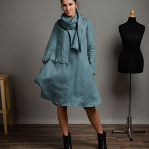 May include: A teal linen dress with long sleeves and a relaxed fit. The dress has a slightly flared skirt and a simple, elegant design. The model is wearing black ankle boots.