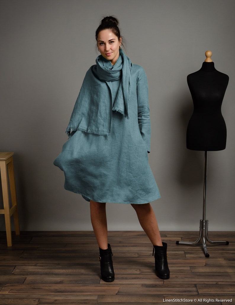 May include: A teal linen dress with long sleeves and a relaxed fit. The dress has a slightly flared skirt and a simple, elegant design. The model is wearing black ankle boots.