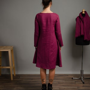 May include: A magenta long-sleeved dress with a round neckline and a flared skirt that falls to the knees. A matching magenta scarf is draped over a dress form standing to the right. The text LinenStitchStore &copy; All Rights Reserved is visible.