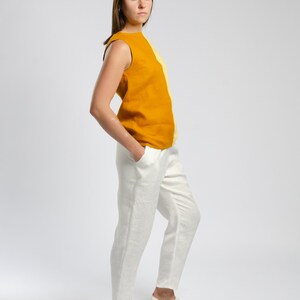 May include: A woman wearing a sleeveless top with a yellow and orange color block design and white linen pants. She is wearing white sneakers with colorful accents.