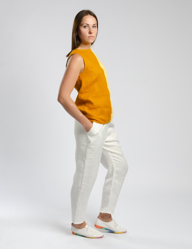 May include: A woman wearing a sleeveless top with a yellow and orange color block design and white linen pants. She is wearing white sneakers with colorful accents.