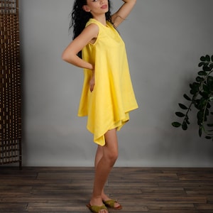 May include: A woman wearing a bright yellow sleeveless linen dress with a flowing, asymmetrical hemline. She is standing on a wood floor with a plant in the background.