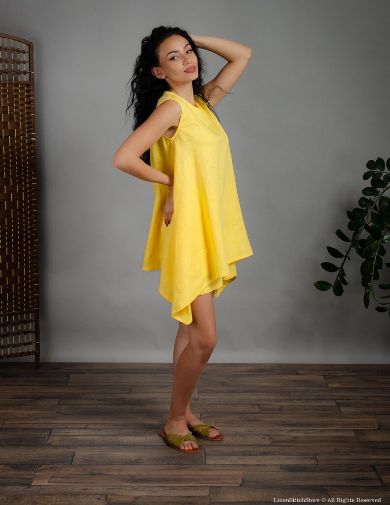 May include: A woman wearing a bright yellow sleeveless linen dress with a flowing, asymmetrical hemline. She is standing on a wood floor with a plant in the background.