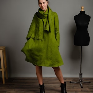 May include: A woman wearing a green linen dress with a matching scarf. She is standing on a wooden floor and wearing black ankle boots.