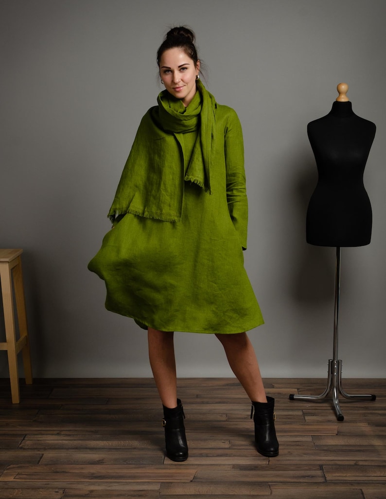 May include: A woman wearing a green linen dress with a matching scarf. She is standing on a wooden floor and wearing black ankle boots.