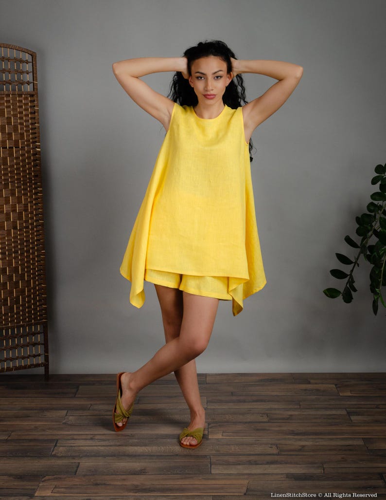 May include: A woman wearing a yellow linen sleeveless tunic top and matching shorts. The tunic has a flowing, asymmetrical hemline. She is wearing green sandals.
