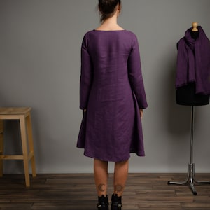 May include: A long-sleeved, purple linen dress with a scoop neck and a slightly flared skirt. The dress is shown on a model with tattoos on her ankles.