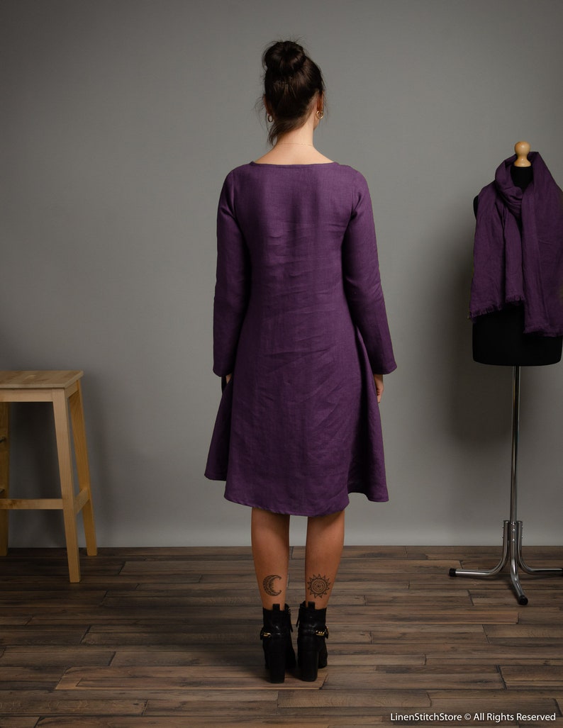 May include: A long-sleeved, purple linen dress with a scoop neck and a slightly flared skirt. The dress is shown on a model with tattoos on her ankles.