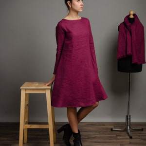 May include: A long-sleeved, knee-length magenta dress with a boat neckline and a flared skirt silhouette. The dress is made of a textured fabric and is displayed alongside a matching magenta scarf draped over a black mannequin form.